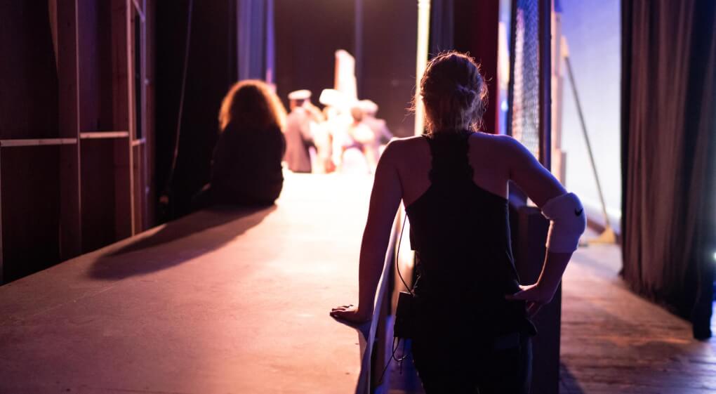 Female student sitting backstage with her back to the camera and stage lights flooding past her